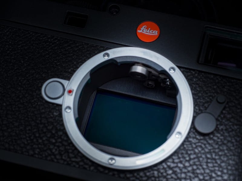 Image 3: Close-up of a Leica camera body without a lens, showing the lens mount and sensor. The red Leica logo is visible at the top, and the camera has a textured black surface.