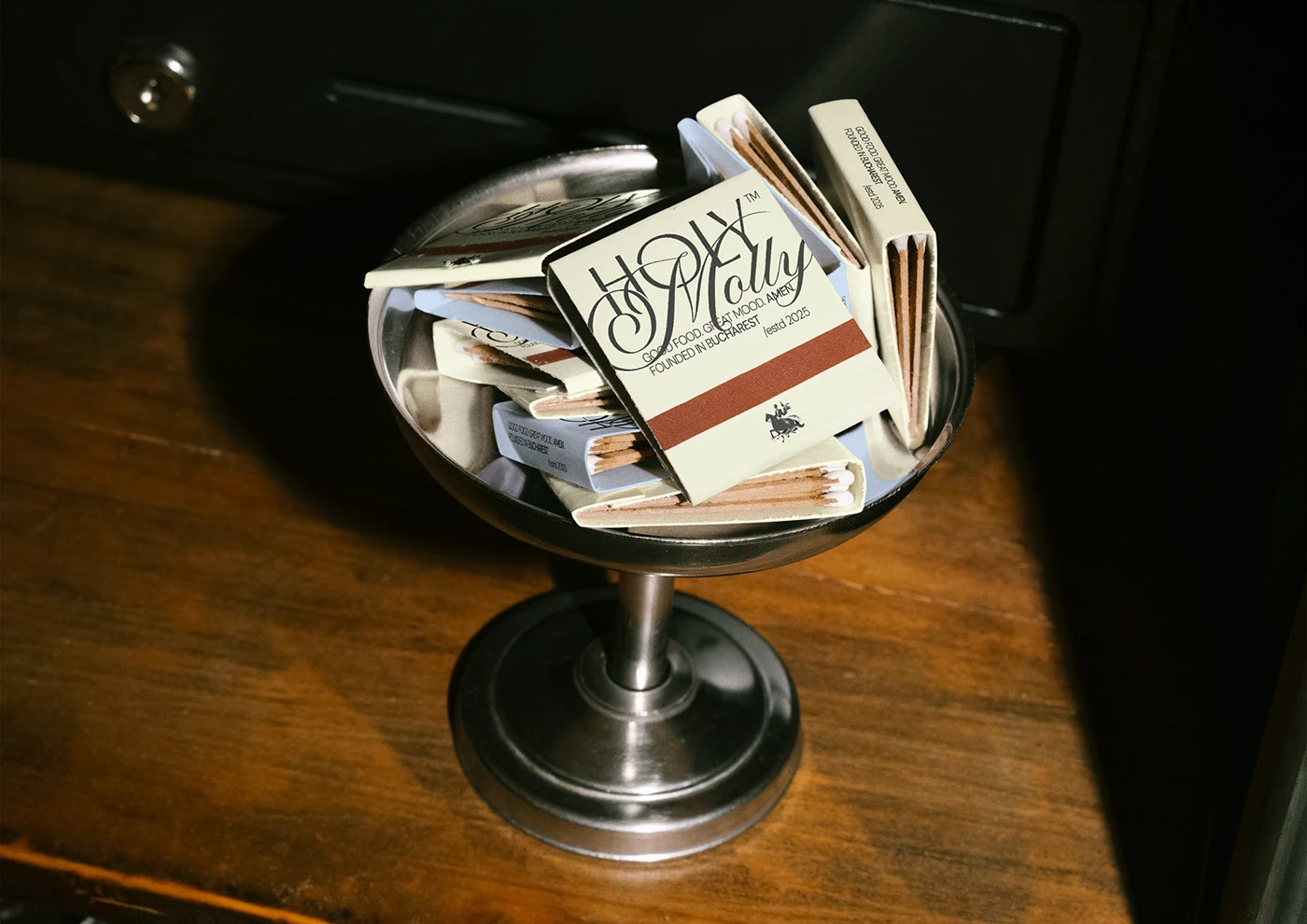 Image 3: Holy Molly branded matchboxes stacked inside a silver coupe glass, black-and-white on cream — restaurant brand identity